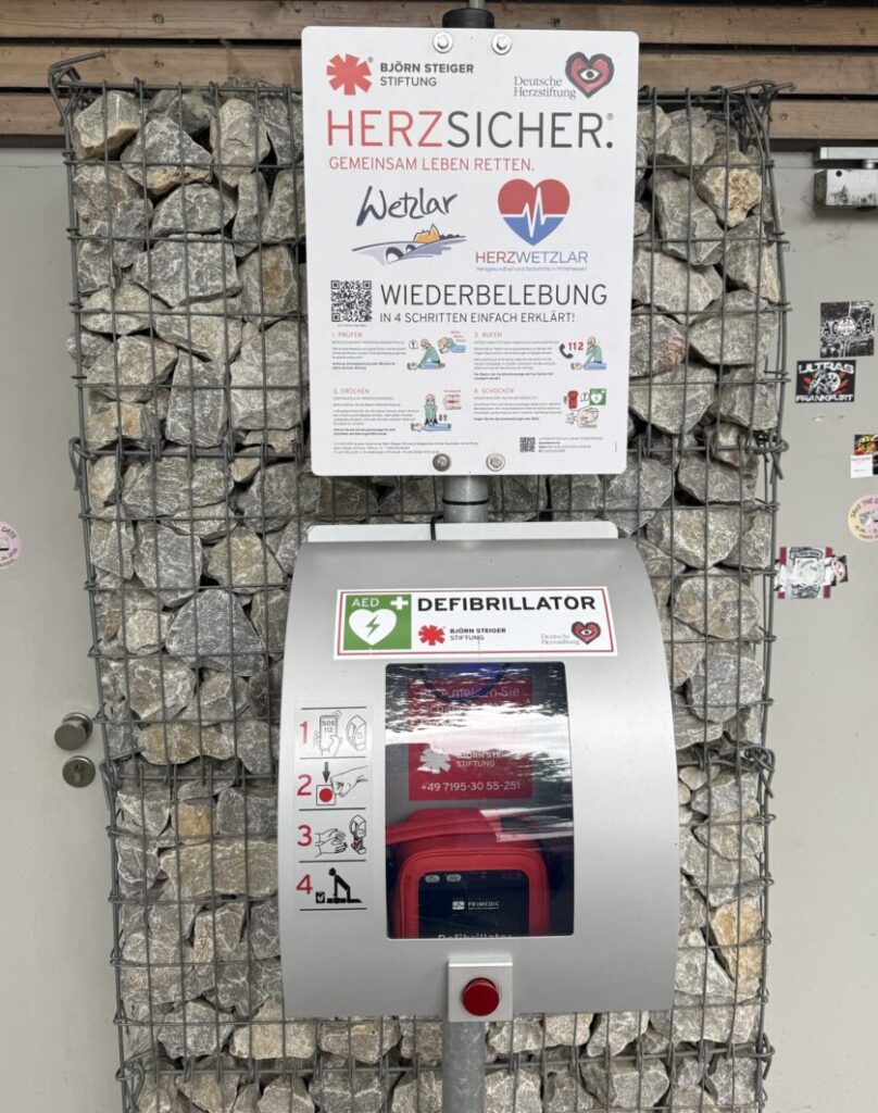 Stahlkasten mit Defibrillator und Schild mit Anleitung zur Wiederbelebung und Titel: "Herzsicher - Gemeinsam Leben retten". Logos von Herz in Wetzlar, Stadt, Wetzlar, Steigerstiftung und Herzstiftung.
